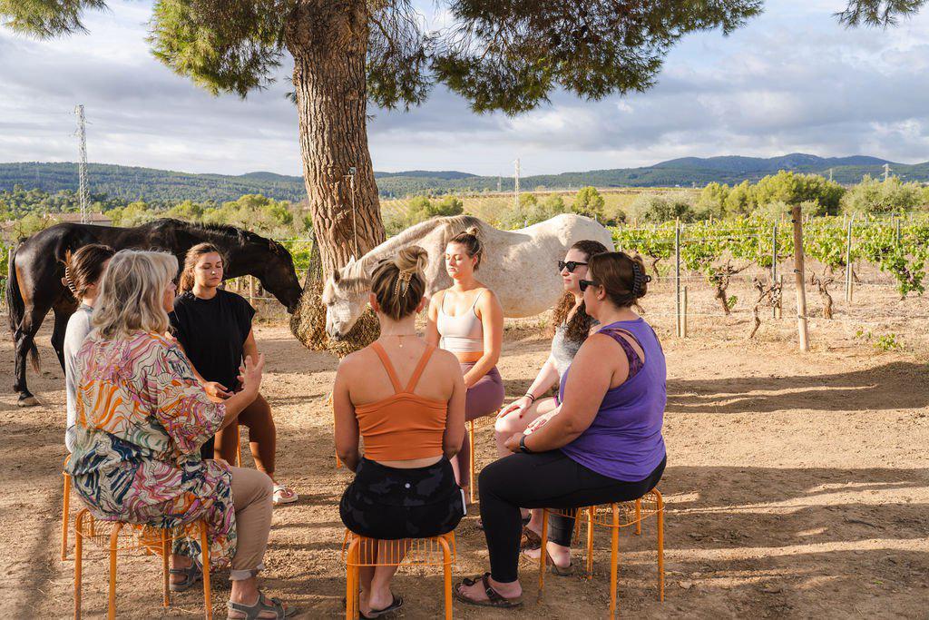 Un grupo de mujeres sentadas en círculo bajo un árbol, con caballos pastando cerca en un pintoresco paisaje de viñedos.